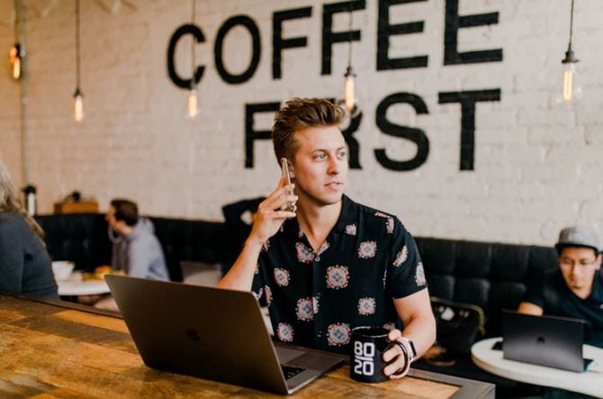 Social Media Manager: Um homem jovem, em uma cafeteria, segurando com o celular no ouvido, com o notebook aberto na mesa ao lado de uma caneca de café.