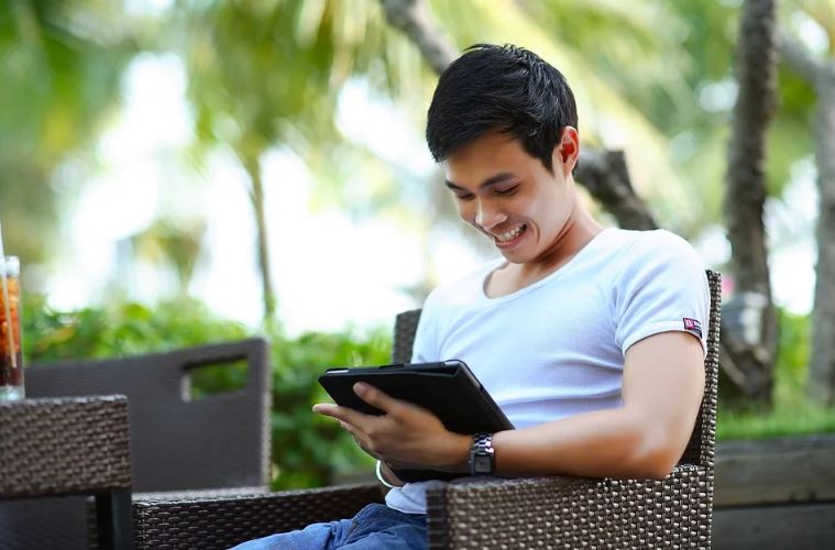 Redes Sociais Novas: imagem de um jovem sentado ao ar livre, segurando um tablete e sorrindo e olhando para a tela.