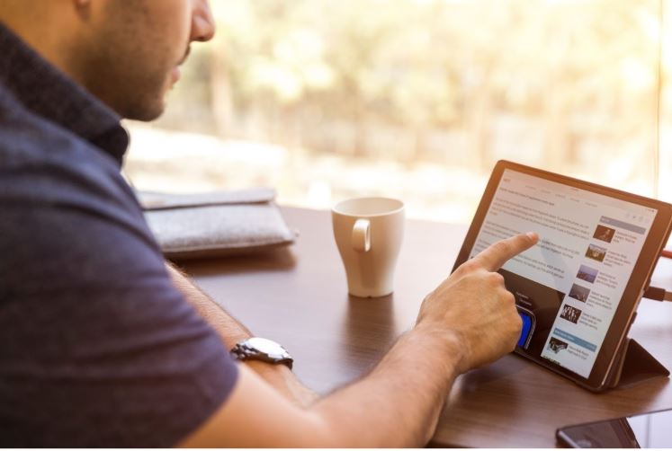 Perfil LinkedIn: imagem de um homem sentado apontando para a tela do tablet com a página do LinkedIn aberta. O tablet está sob uma mesa de madeira com uma caneca branca de cerâmica e um celular ao lado.