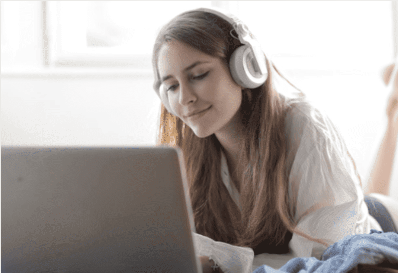 Como fazer podcast: uma jovem branca está deitada na cama e olha, sorrindo, para a tela de um computador enquanto escuta algo em um fone de ouvido.
