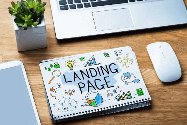 Criar landing page: imagem de uma página de caderno, escrito landing page, sob uma mesa de madeira ao lado de um notebook e uma planta