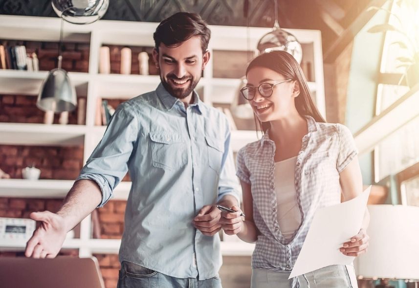 tecnicas de copywriting: imagem de um homem e uma mulher, ambos em pé, conversando, ela segura um papel, enquanto ele aponta com a mão para a tela de um notebook